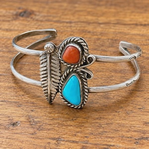 May include: A silver cuff bracelet featuring a central design with a red stone and a blue stone, each framed by twisted metal wire. To the left of the stones is a silver feather motif with etched lines and a small circular accent.