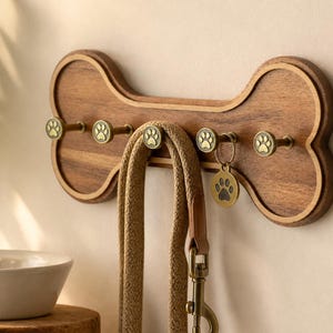 Könnte beinhalten: Ein wandmontierter Leinenhalter aus Holz in Knochenform mit fünf messingfarbenen Haken, die jeweils mit einem Pfotenabdruck-Design versehen sind. Eine beige gewebte Leine hängt am mittleren Haken und ein runder Anhänger mit Pfotenabdruck.