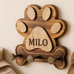 May include: A wooden wall-mounted hook rack shaped like a paw print and a bone. The central paw pad is engraved with the name MILO. It features three bronze-toned metal hooks, each topped with a small paw print design, and a measuring tape nearby.
