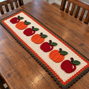 Puede incluir: Un camino de mesa rectangular de ganchillo con un panel central de color blanco roto decorado con un patrón alterno de manzanas rojas y calabazas naranjas, cada una con hojas verdes, bordeado por bordes festoneados naranjas y marrones.