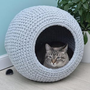 Puede incluir: Una cama esférica para mascotas tejida a ganchillo en color gris claro con una gran abertura circular. El exterior está hecho de hilo grueso y texturizado con un patrón de punto consistente. Dentro de la estructura redonda, descansa un gato atigrado.