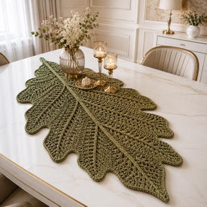 May include: A large, olive green crochet table runner shaped like an elongated oak leaf with intricate, open-work stitching that mimics natural leaf veins. The textile piece features a central spine and scalloped edges, providing a textured surface.