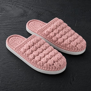 May include: A pair of dusty pink slippers featuring a textured, crocheted upper with a raised, repeating puff stitch pattern. The slippers have a solid, flat white sole and an open-back design, showcasing a consistent yarn construction throughout.