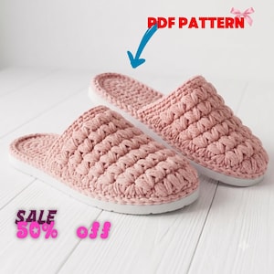May include: A pair of dusty pink crochet slippers with a textured puff stitch upper and a solid white sole. The image includes the text PDF PATTERN with a blue arrow pointing to the slippers and the text SALE 50% off in pink lettering.