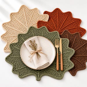 May include: A set of four leaf-shaped crochet placemats in beige, rust orange, olive green, and dark brown. A round white plate sits on the green placemat, topped with a folded beige napkin tied with twine and a small dried sprig, plus a fork and knife.