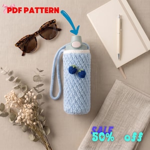 May include: A light blue crocheted water bottle cozy featuring a textured stitch pattern, a matching carrying strap, and two small dark blue berry appliques. The cozy is shown with brown tortoiseshell sunglasses, a white notebook, and a pen.