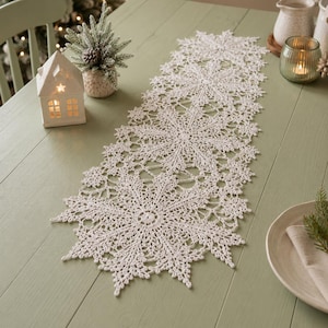 May include: A long, off-white crochet table runner featuring a repeating intricate snowflake pattern with pointed edges. The textile is crafted with fine thread, showcasing a delicate, openwork design that spans the length of the table surface.