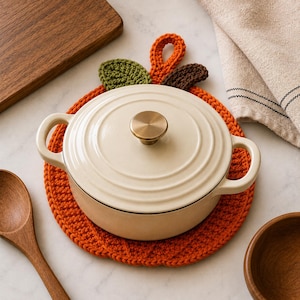 May include: A cream-coloured enamelled cast iron Dutch oven with a round lid and a metallic knob, resting on a round, orange, crocheted pumpkin-shaped trivet featuring a green leaf, a brown leaf, and an orange stem loop at the top.
