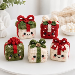 Op de afbeelding: Vijf vierkante gehaakte cadeau-doosjes met lachende gezichtjes, zwarte ogen en roze wangen. Elke doos heeft een gehaakte strik bovenop, sommige met groene blaadjes of een houten knoop. De kleuren zijn crème, rood en olijfgroen met sneeuwvlokpatronen.