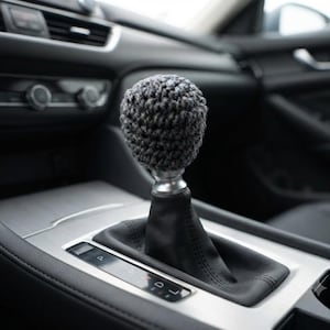 May include: A spherical, dark gray crocheted cover fitted over a car gear shift knob. The cover features a textured, looped yarn pattern and sits atop a silver metal base, which connects to a black leather gear shift boot in a vehicle interior.
