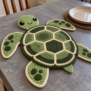 May include: A crochet turtle-shaped mat featuring a shell with dark green, light green, and cream-coloured hexagonal patterns. The head has black and white eyes and a stitched smile, while the four flippers display dark green circular accents.