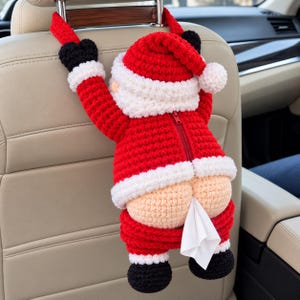 May include: A crocheted Santa Claus tissue holder hanging from a car headrest. The figure features a red suit with white trim, black boots, and mittens. A zipper runs down the center of the jacket, and a white tissue is dispensed from the rear.