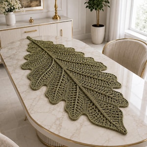 Peut inclure: Un grand chemin de table en crochet vert olive en forme de feuille de chêne allongée. Le textile présente un motif de mailles ajourées détaillé qui imite les nervures et les bords lobés d'une feuille naturelle, s'étendant sur la table.