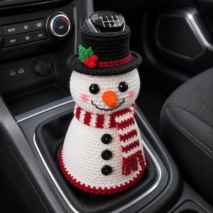 May include: A white crocheted snowman gear shift cover featuring a black top hat with green holly and red berries, a bright orange carrot nose, pink cheeks, three black buttons, and a red and white striped scarf wrapped around the body.