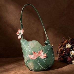 May include: A green shoulder bag with a curved, leaf-like silhouette featuring embroidered pink lotus flower details on the front and a single thin strap. The bag has a smooth finish with white line accents that mimic the veins of a leaf.