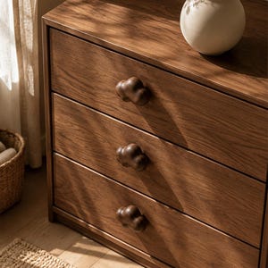 Peut inclure: Une commode en bois dot&eacute;e de trois tiroirs horizontaux avec des boutons en bois fonc&eacute; en forme de nuage. Le meuble pr&eacute;sente un grain de bois naturel visible et une finition lisse. Un vase en c&eacute;ramique blanc cass&eacute; est pos&eacute; dessus.