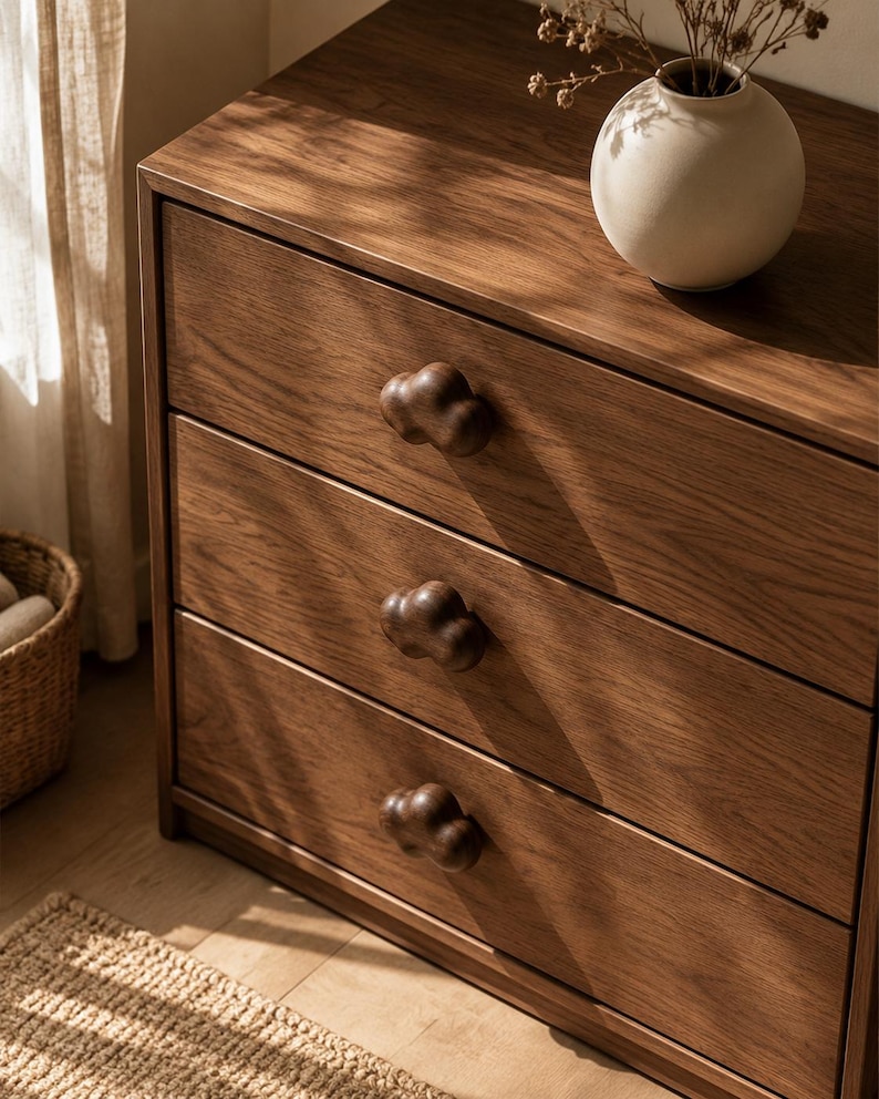 Peut inclure: Une commode en bois dot&eacute;e de trois tiroirs horizontaux avec des boutons en bois fonc&eacute; en forme de nuage. Le meuble pr&eacute;sente un grain de bois naturel visible et une finition lisse. Un vase en c&eacute;ramique blanc cass&eacute; est pos&eacute; dessus.