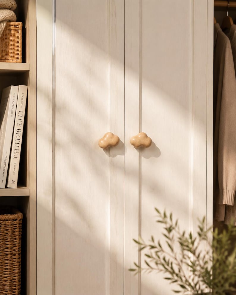 Peut inclure: Deux boutons de placard en bois clair en forme de nuages sont fix&eacute;s au centre d'une paire de portes de placard verticales &agrave; panneaux blanc cass&eacute;. Les boutons ont une finition lisse et arrondie et sont plac&eacute;s c&ocirc;te &agrave; c&ocirc;te sur les portes.