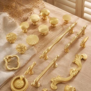 May include: A collection of gold-toned metal cabinet hardware including various drawer pulls, knobs, and handles. The set features smooth round knobs, floral-shaped knobs, ring pulls, and elongated handles with ornate, curved, and straight designs.