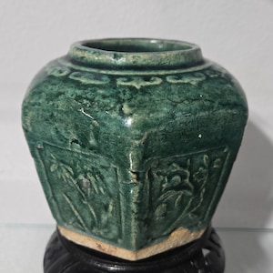 Peut inclure: Un pot en céramique hexagonal avec une finition émaillée vert foncé craquelé. L'extérieur présente des motifs floraux et botaniques en relief sur chaque face, avec une large ouverture circulaire au sommet et une base non émaillée.
