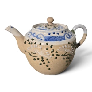 May include: A ceramic teapot with a beige textured body featuring raised green dot accents and white floral patterns. The upper section is decorated with blue painted swirls and dots, and it includes a matching lid with a small knob handle.