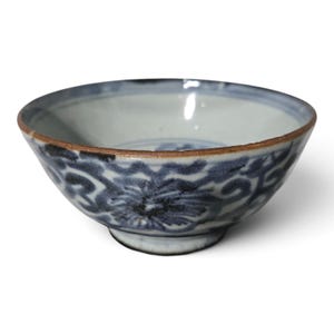 May include: A ceramic bowl featuring a white base with blue floral patterns painted around the exterior. The bowl has a wide, open mouth and a distinct brown rim along the top edge, with a circular footed base at the bottom.