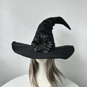 May include: A black pointed hat with a wide brim and a tall, slightly curved crown. The hat features a decorative black flower embellishment centered on the front band. It is displayed on a mannequin head with long, straight, light gray hair.