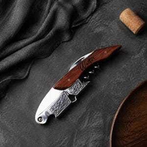 May include: A folding wine opener featuring a dark wood handle with the name Walter engraved in script. The tool includes a silver-coloured metal lever with an embossed leaf pattern, a metal worm, and a small blade, all integrated into a compact design.
