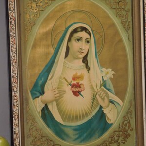 May include: Framed religious print featuring a depiction of the Virgin Mary. She is wearing a blue veil and holding a white lily. Her chest displays a heart pierced by a sword. The artwork is set within an oval frame, surrounded by decorative elements.