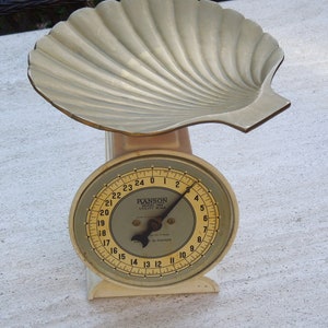 May include: A vintage white kitchen scale with a gold-colored shell-shaped top and a round dial with black markings. The dial reads from 0 to 24 and is labeled "HANSON UTILITY SCALE 25 POUNDS".