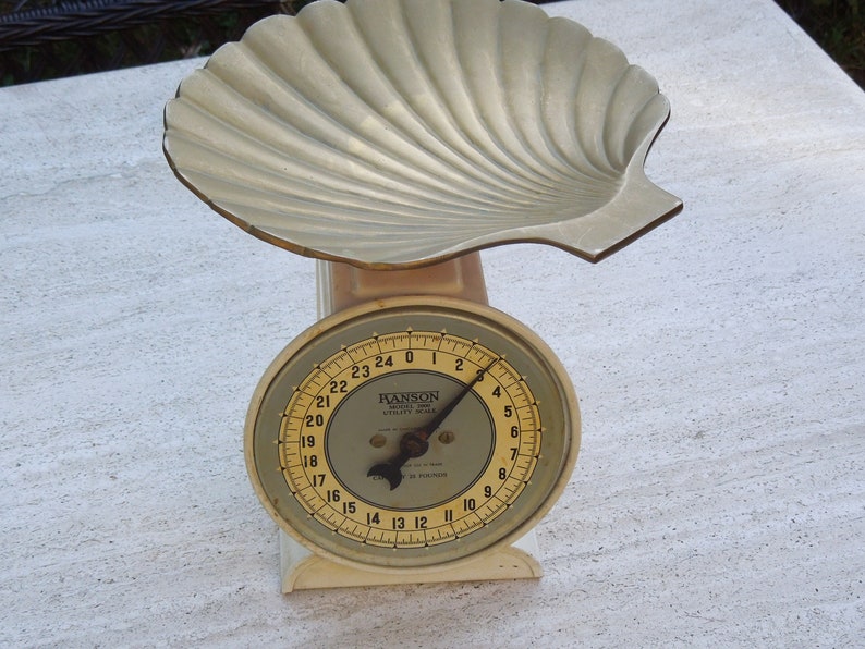 May include: A vintage white kitchen scale with a gold-colored shell-shaped top and a round dial with black markings. The dial reads from 0 to 24 and is labeled "HANSON UTILITY SCALE 25 POUNDS".