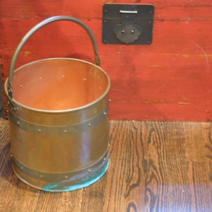 May include: A copper bucket with a brass handle and bands. The bucket is sitting on a wooden floor.