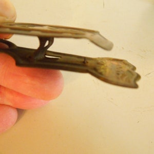 May include: An antique metal clothes peg with a spring mechanism. The peg is a dark, aged colour with a decorative design on the top and a textured grip on the bottom. The peg is open, ready to be used.
