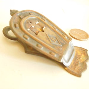 May include: Vintage brass horseshoe-shaped paper clip. The clip features an intricate design with a diamond motif and a small loop. A penny is placed nearby for scale. The clip is approximately 7 cm long.