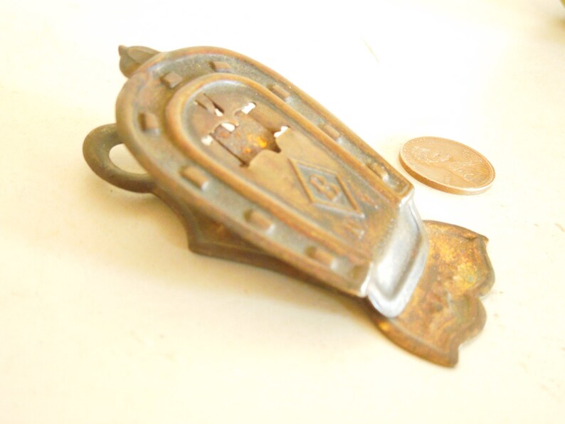 May include: Vintage brass horseshoe-shaped paper clip. The clip features an intricate design with a diamond motif and a small loop. A penny is placed nearby for scale. The clip is approximately 7 cm long.