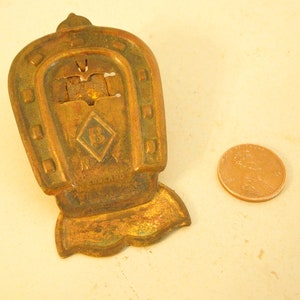 May include: A vintage brass horseshoe-shaped object with a diamond emblem and the letter "B". It has a rectangular base and is next to a penny. The text "ENGLAND" is visible. The object is likely a charm.