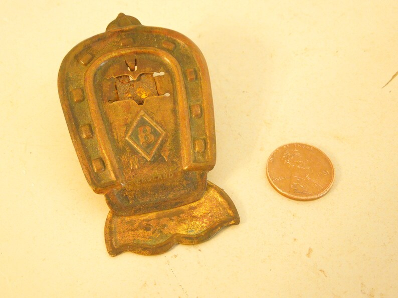 May include: A vintage brass horseshoe-shaped object with a diamond emblem and the letter "B". It has a rectangular base and is next to a penny. The text "ENGLAND" is visible. The object is likely a charm.