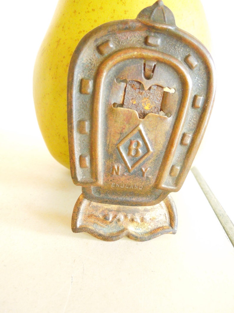 May include: A vintage bronze horseshoe-shaped bottle opener with the text "N.Y. ENGLAND" and a diamond with the letter "B" inside. The opener has a decorative top and a small base. A yellow pear is in the background.