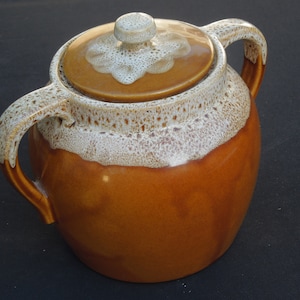 May include: A brown ceramic crock with a white glaze and two handles. The lid has a white knob and a white design.