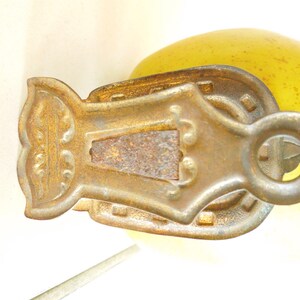 May include: An antique brass-coloured pulley with an ornate design. The pulley has a central rectangular section with a rusted appearance, surrounded by decorative elements and a circular ring for attachment. The background is a soft yellow.