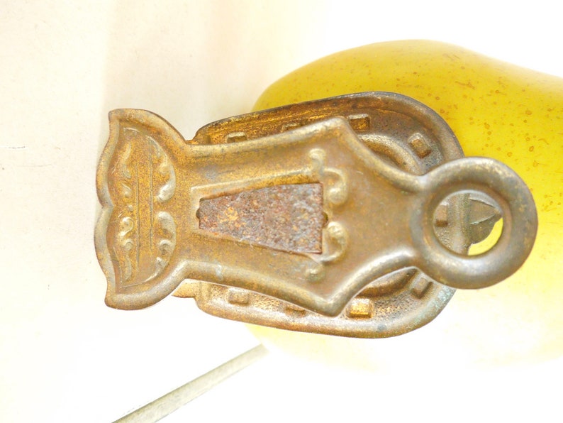 May include: An antique brass-coloured pulley with an ornate design. The pulley has a central rectangular section with a rusted appearance, surrounded by decorative elements and a circular ring for attachment. The background is a soft yellow.