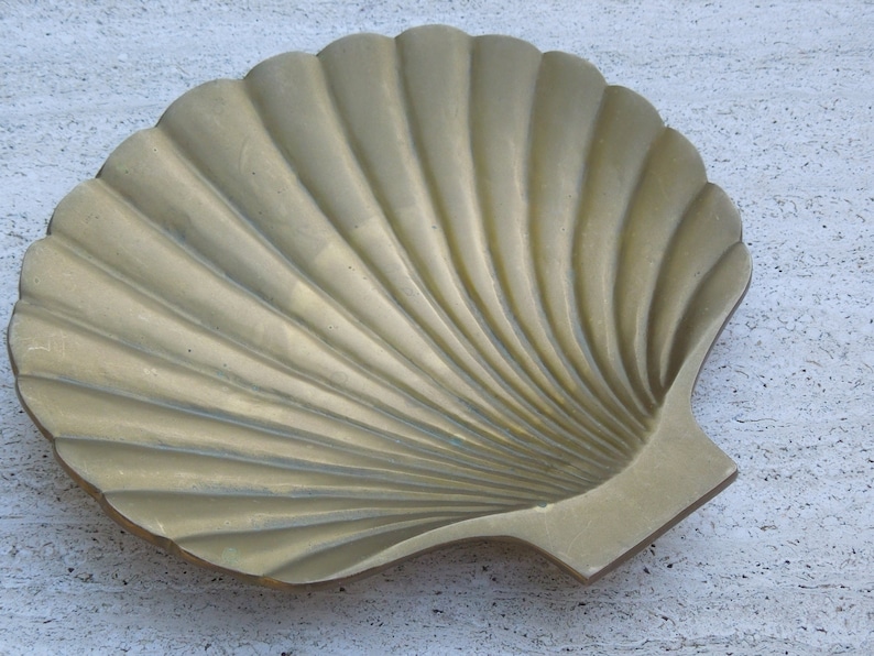 May include: A gold-colored metal seashell-shaped dish. The dish is made to look like a scallop shell with a ribbed texture.