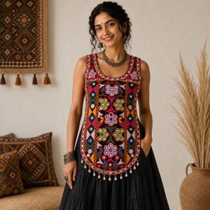 Könnte beinhalten: Eine ärmellose Tunika mit einem lebendigen, mehrfarbigen Stickmuster aus kreisförmigen Spiegeln, geometrischen Formen und Blumenmotiven auf schwarzem Grund, abgeschlossen mit einer Reihe kleiner, weißer Muscheln am abgerundeten Saum.