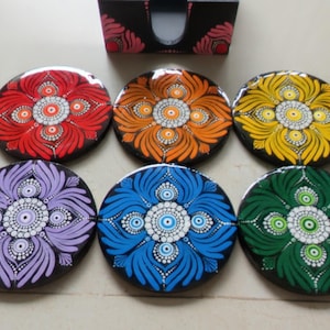 May include: A set of six circular coasters featuring intricate dot-painted mandala patterns in red, orange, yellow, purple, blue, and green, arranged with a matching black storage holder that displays pink floral accents at the top corners.