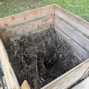May include: A square wooden compost bin constructed from horizontal slats and wire mesh panels. The interior is filled with a dense pile of dry brown twigs, organic plant debris, and dark soil matter, showing a collection of garden waste.
