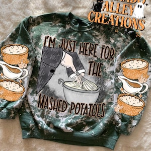 Peut inclure: Un sweat-shirt à col rond vert foncé délavé avec un motif représentant une main fouettant de la purée dans un bol. Le texte indique I'M JUST HERE FOR THE MASHED POTATOES. Les manches sont ornées d'illustrations de bols de purée et de saucières.