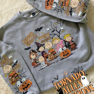May include: A heather gray crewneck sweatshirt featuring a Peanuts character Halloween graphic. The design includes Snoopy, Charlie Brown, and friends in costumes with the text Good Grief It's Spooky Season. The sleeves also feature matching graphics.