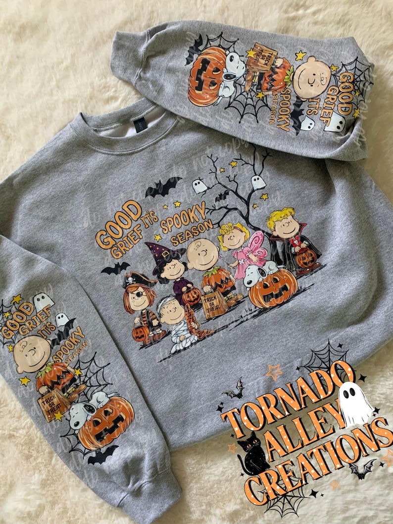 May include: A heather gray crewneck sweatshirt featuring a Peanuts character Halloween graphic. The design includes Snoopy, Charlie Brown, and friends in costumes with the text Good Grief It's Spooky Season. The sleeves also feature matching graphics.
