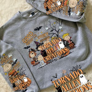 May include: A heather gray crewneck sweatshirt featuring a Halloween-themed graphic with cartoon characters in costumes, pumpkins, ghosts, bats, and spiderwebs. The text reads "GOOD GRIEF IT'S SPOOKY SEASON" and "TORNADO ALLEY CREATIONS" on the front and sleeves.