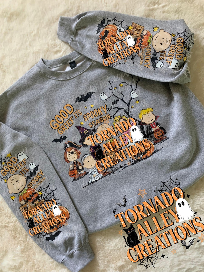 May include: A heather gray crewneck sweatshirt featuring a Halloween-themed graphic with cartoon characters in costumes, pumpkins, ghosts, bats, and spiderwebs. The text reads "GOOD GRIEF IT'S SPOOKY SEASON" and "TORNADO ALLEY CREATIONS" on the front and sleeves.